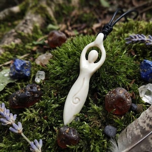 Könnte beinhalten: Elfenbeinfarbene Anhänger-Halskette mit einer stilisierten weiblichen Figur, die an einer schwarzen Kordel hängt. Der Anhänger ist von Moos, Edelsteinen und einer Feder umgeben. Ein spirituelles oder böhmisches Accessoire.