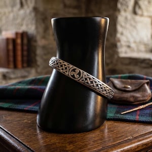 May include: A silver Celtic knot bracelet on a black display form. The bracelet has an intricate knot design. A small leather pouch and a decorative pin are on a wooden surface, with a plaid textile in the background. The setting suggests Scottish heritage.
