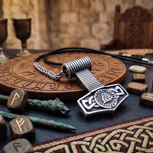 Könnte beinhalten: Eine silberfarbene Halskette mit einem Hammeranhänger, inspiriert von der nordischen Mythologie. Der Anhänger ist an einem schwarzen Band und einer silbernen Kette befestigt. Holzrunen und dekorative Elemente sind ebenfalls im Bild zu sehen.