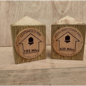 May include: Two wooden trail markers with circular plaques. One reads "Cotswold Way" with "102 Miles" and dates. The other says "South West Coast Path" with "630 Miles" and dates. The markers are on a light wood surface.
