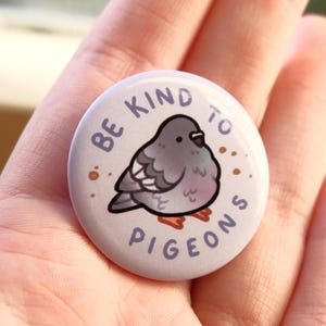 May include: A round, light purple button with the text "BE KIND TO PIGEONS" encircling a cartoon pigeon illustration. The pigeon is gray with pink accents and brown speckles. The button is held in a hand.