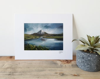 May include: An original landscape painting of a mountain reflected in a lake, framed in a white mat. The artwork features a snow-capped mountain, green hills, and a cloudy sky. The painting is displayed on a wooden surface.