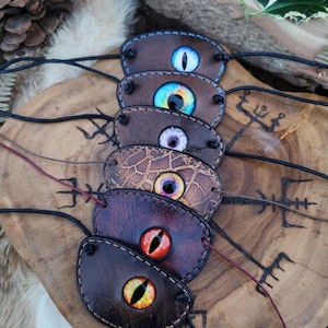 Peut inclure: Plusieurs cache-œil en cuir faits à la main, chacun avec un design d'œil coloré unique. Les patchs bruns ont des cordons noirs et les yeux sont de différentes couleurs, dont le bleu, le violet et l'orange. Les patchs sont présentés sur une surface en bois.