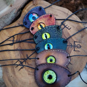 以下が含まれることがあります： リアルでカラフルな目のデザインが施された、手作りの眼帯のコレクション。各パッチは紫、オレンジ、緑、茶色など異なる色で、黒いコードが付いています。木製の表面に展示されています。