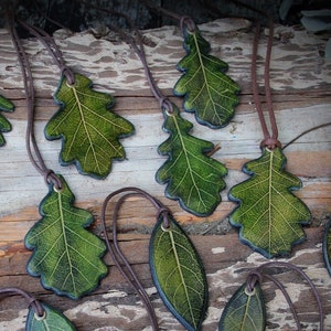 Può includere: Una collezione di ciondoli a forma di foglia con cordini marroni. I ciondoli sono di varie tonalità di verde, con venature dettagliate, e sono disposti su una superficie di legno invecchiato. Il tema generale è la gioielleria ispirata alla natura.