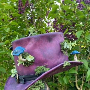May include: A deep purple top hat with a black band and buckle, adorned with blue mushroom ornaments, green moss, and small green leaves. The hat is set against a backdrop of green foliage and purple flowers.