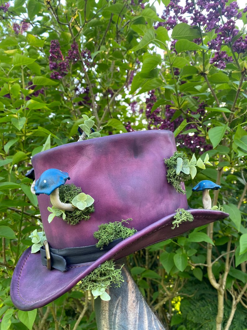 May include: A deep purple top hat with a black band and buckle, adorned with blue mushroom ornaments, green moss, and small green leaves. The hat is set against a backdrop of green foliage and purple flowers.