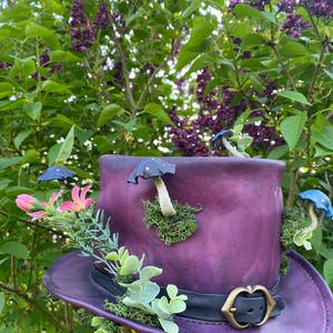 May include: A purple leather hat with a wide brim, adorned with miniature blue and white mushrooms, pink flowers, and green foliage. The hat has a black band with a gold-coloured buckle. The background features green leaves and purple flowers.