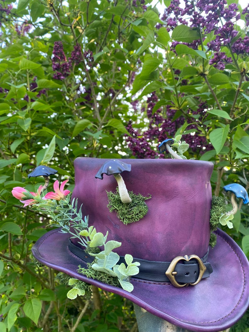 May include: A purple leather hat with a wide brim, adorned with miniature blue and white mushrooms, pink flowers, and green foliage. The hat has a black band with a gold-coloured buckle. The background features green leaves and purple flowers.