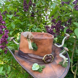 May include: A brown leather cowboy hat decorated with faux mushrooms, moss, and a brass buckle. The hat has a wide brim and a dark brown leather band. The mushrooms are various sizes and colors, adding a whimsical touch to the hat.