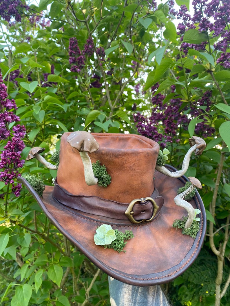 May include: A brown leather cowboy hat decorated with faux mushrooms, moss, and a brass buckle. The hat has a wide brim and a dark brown leather band. The mushrooms are various sizes and colors, adding a whimsical touch to the hat.