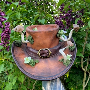 May include: A brown leather cowboy hat decorated with faux mushrooms, moss, and small flowers. The hat features a decorative buckle and is set against a backdrop of green foliage and purple flowers.