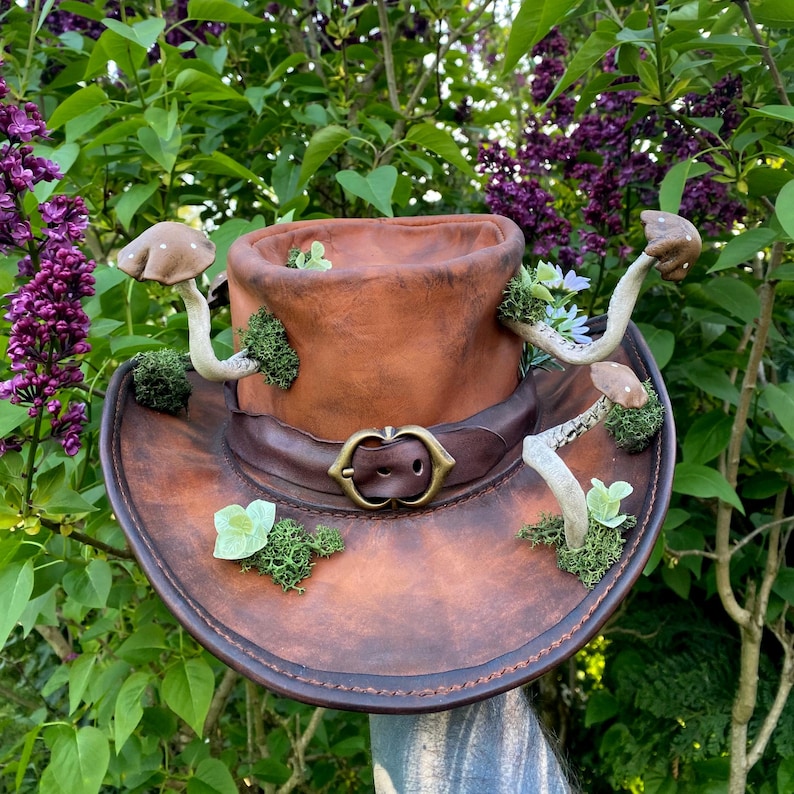 May include: A brown leather cowboy hat decorated with faux mushrooms, moss, and small flowers. The hat features a decorative buckle and is set against a backdrop of green foliage and purple flowers.