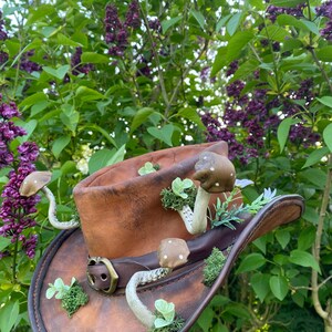 May include: A brown leather cowboy hat adorned with whimsical mushroom and plant accents. The hat features a decorative buckle and is set against a backdrop of lush green foliage and purple flowers, creating a unique, nature-inspired accessory.