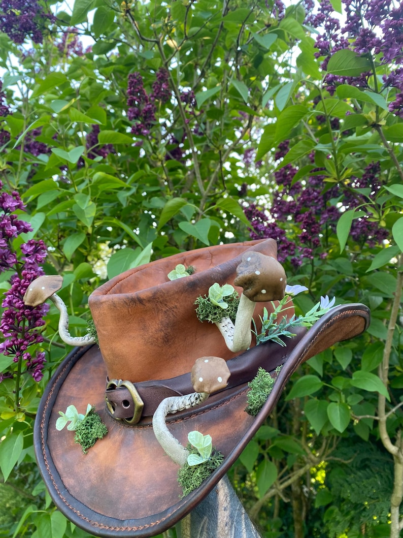 May include: A brown leather cowboy hat adorned with whimsical mushroom and plant accents. The hat features a decorative buckle and is set against a backdrop of lush green foliage and purple flowers, creating a unique, nature-inspired accessory.