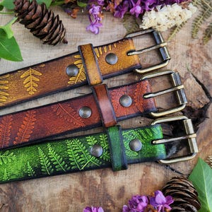 Op de afbeelding: Vier leren riemen met bladeren in bruin, rood en groen. Elke riem heeft een metalen gesp en is versierd met een varenpatroon. De riemen worden gepresenteerd op een houten oppervlak met paarse bloemen en dennenappels.