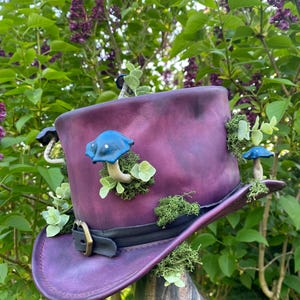 May include: A deep purple top hat featuring a black band and buckle, decorated with blue mushroom details and green moss. The hat is embellished with small green plants. A whimsical accessory.