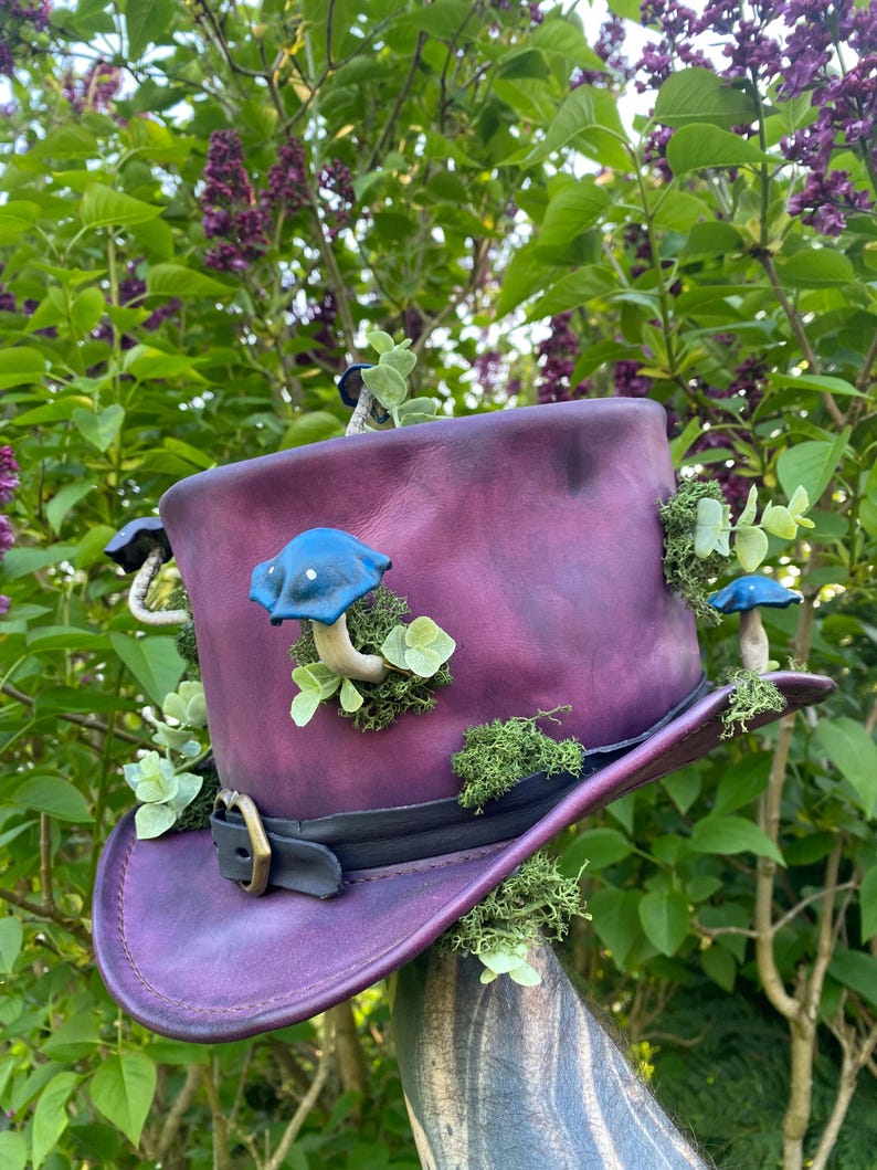 May include: A deep purple top hat featuring a black band and buckle, decorated with blue mushroom details and green moss. The hat is embellished with small green plants. A whimsical accessory.