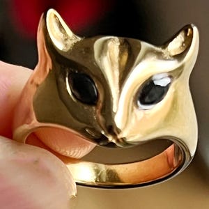 Peut inclure: Une bague dorée en forme de tête de chat. La bague présente des yeux noirs et des oreilles pointues. La bague est tenue entre les doigts, mettant en valeur sa surface polie et réfléchissante.