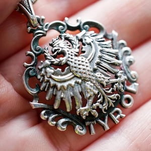 Könnte beinhalten: Silberanhänger mit detailliertem Wappenadler-Design. Der Adler befindet sich in einem verzierten, durchbrochenen Rahmen. Der Anhänger hat eine strukturierte, antike Oberfläche, und das Wort "TYR" ist sichtbar.