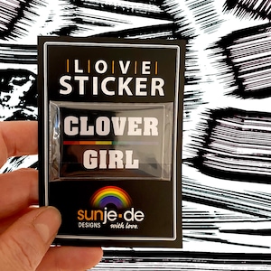 May include: A black and white sticker with the text "Clover Girl" in white letters. The sticker has a rainbow stripe above the text. The sticker is packaged in a black card with the text "I Love Sticker" in white letters. The card also has the text "sunje.de Designs with love." in white letters.