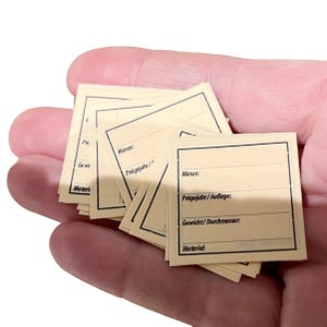 May include: A handful of small, rectangular, cream-colored labels with black borders and lines for writing. The labels have German text, including "Münze" and "Prägejahr/Auflage:" They are designed for organizing or labeling items.