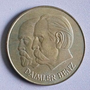 May include: A bronze-colored coin featuring two side profiles of men with mustaches and the text "DAIMLER BENZ" in a circular arrangement. The coin has a slightly worn appearance.