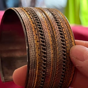Könnte beinhalten: Ein breites Armband mit einem mehrschichtigen Design aus abwechselnden Bändern in metallischen Gold-, Kupfer- und Silbertönen. Die Oberfläche zeigt eine komplexe Textur, die aus feinen Kettenmustern und horizontalen Reihen von Metallsegmenten besteht.