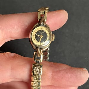 May include: Vintage gold-toned wristwatch with a black face and gold hands. The watch features the brand name "Stowa" on the face. The watch band is a gold-toned metal with a textured design. The watch is held by a person's fingers.