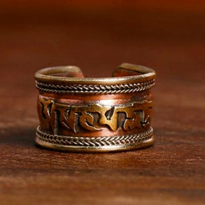 Puede incluir: Un anillo de tono cobrizo con un diseño abierto. El anillo presenta una banda con letras grabadas y detalles trenzados. El anillo tiene una estética vintage y está sobre un fondo marrón cálido.