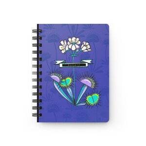 Puede incluir: Cuaderno de espiral con una cubierta morada que presenta una ilustración caprichosa. El diseño incluye una Venus atrapamoscas, flores blancas y la palabra "hangry" en una pancarta. El cuaderno tiene una encuadernación en espiral negra.