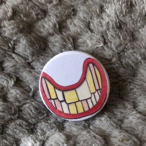 May include: A white button with a red outline and a yellow and pink cartoon mouth with teeth.