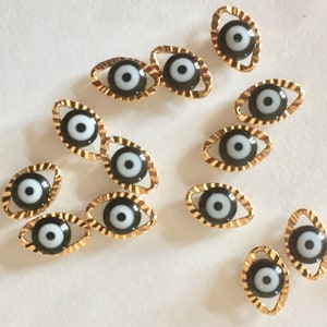 May include: A collection of gold-colored evil eye charms with black and white enamel. The charms are oval-shaped and have a detailed, textured edge.