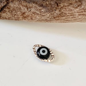May include: A small, silver charm with a black and white evil eye design. The charm is oval shaped and has a textured edge.