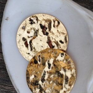 Puede incluir: Dos discos de metal de color dorado con textura martillada, presentados en un plato de cerámica blanca. El plato está sobre una superficie de madera oscura.