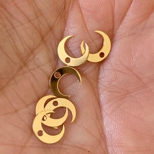 Op de afbeelding: Een groep van vijf gouden halve maan bedels met een klein gaatje in elk. De bedels liggen op de hand van een persoon.