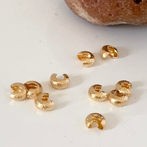 Könnte beinhalten: Nahaufnahme von zehn goldfarbenen Crimpperlen. Die Perlen sind klein und rund, mit einer strukturierten Oberfläche. Sie sind in einem verstreuten Muster auf einer weißen Oberfläche angeordnet.