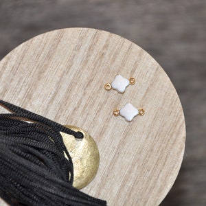 Puede incluir: Dos abalorios blancos en forma de tr&eacute;bol con detalles dorados sobre una superficie de madera. Los abalorios tienen un agujero en el centro para sujetarlos a joyas.