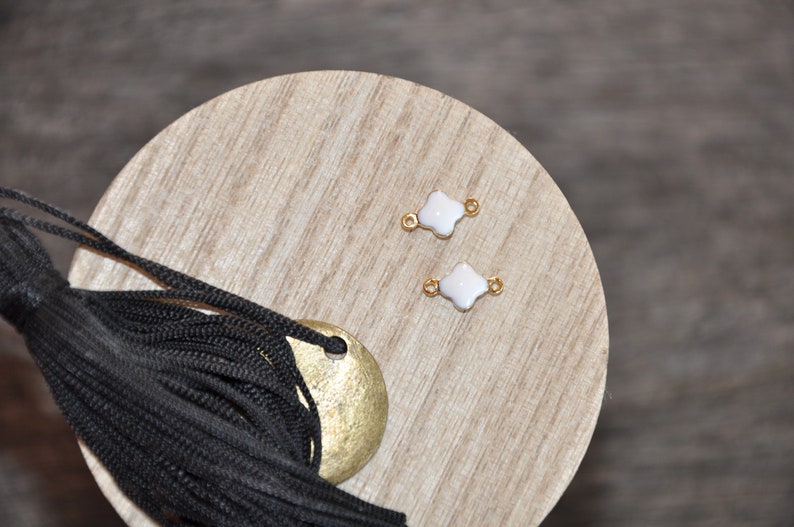 Puede incluir: Dos abalorios blancos en forma de tr&eacute;bol con detalles dorados sobre una superficie de madera. Los abalorios tienen un agujero en el centro para sujetarlos a joyas.