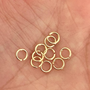 Op de afbeelding: Een verzameling kleine, goudkleurige verbindingsringen, waarschijnlijk voor het maken van sieraden of knutselen. De ringen zijn rond met een kleine opening en worden in een hand gehouden. De ringen hebben een diameter van ongeveer 0,6 cm.