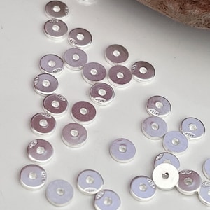 Puede incluir: Un grupo de cuentas espaciadoras de metal de color plateado. Las cuentas son redondas con un agujero en el centro. Las cuentas están dispersas sobre una superficie blanca.