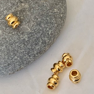 以下が含まれることがあります： テクスチャ加工が施された、隆起したデザインの金色の金属ビーズ。ビーズは白い表面に一列に並んでいます。