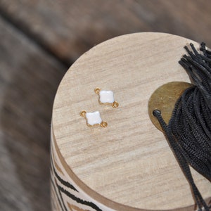 Puede incluir: Dos abalorios conectores de esmalte blanco con detalles dorados. Los abalorios tienen forma de tr&eacute;bol de cuatro hojas y dos peque&ntilde;os bucles a cada lado. Los abalorios est&aacute;n sobre una superficie de madera con un patr&oacute;n geom&eacute;trico blanco y negro.