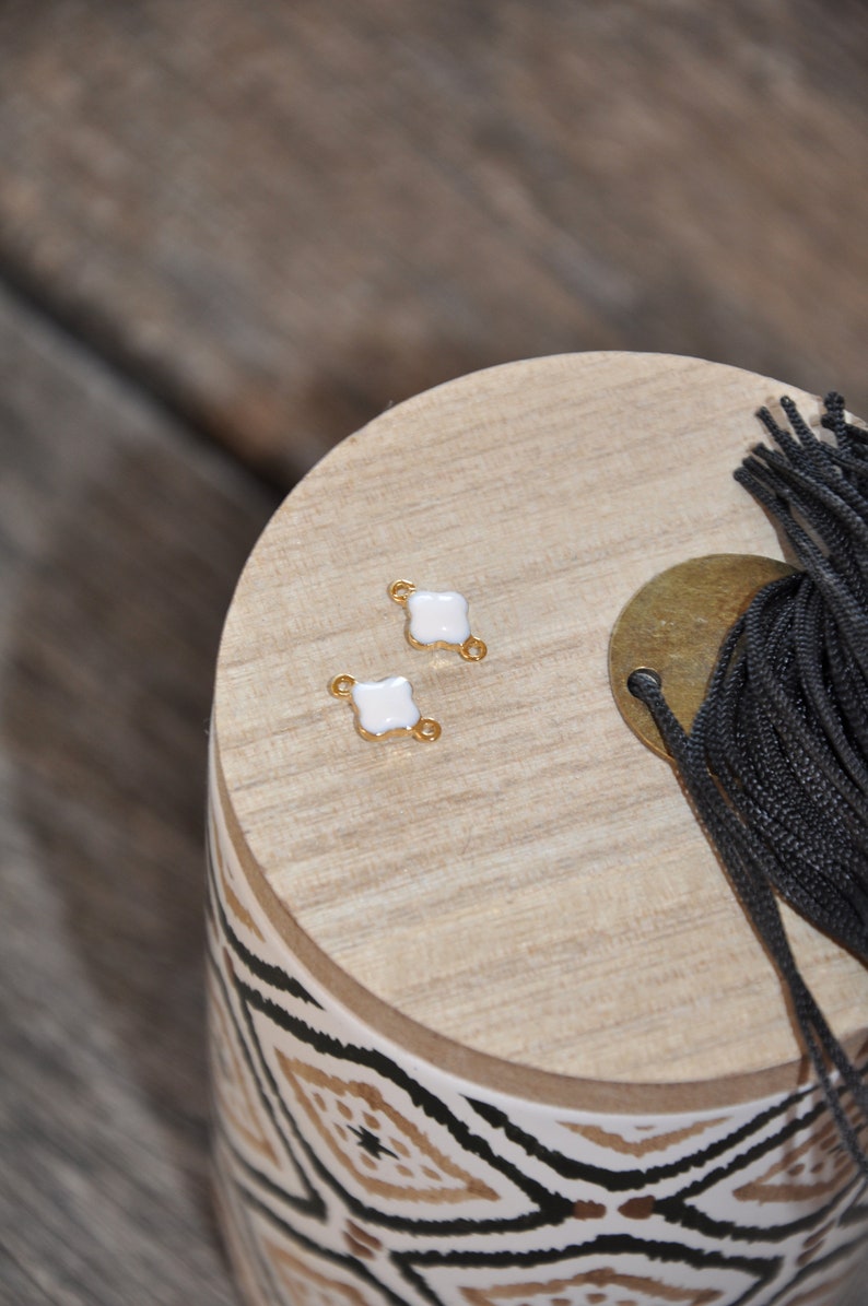 Puede incluir: Dos abalorios conectores de esmalte blanco con detalles dorados. Los abalorios tienen forma de tr&eacute;bol de cuatro hojas y dos peque&ntilde;os bucles a cada lado. Los abalorios est&aacute;n sobre una superficie de madera con un patr&oacute;n geom&eacute;trico blanco y negro.