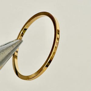 May include: A thin, gold-colored ring held by a pair of silver-colored tweezers. The ring has a simple, minimalist design and appears to be made of metal. The ring is circular and has a smooth, polished surface.