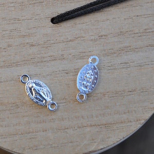 Op de afbeelding: Twee zilveren ovale bedels met een religieus beeld in het midden. De bedels hebben aan elke kant twee lussen.