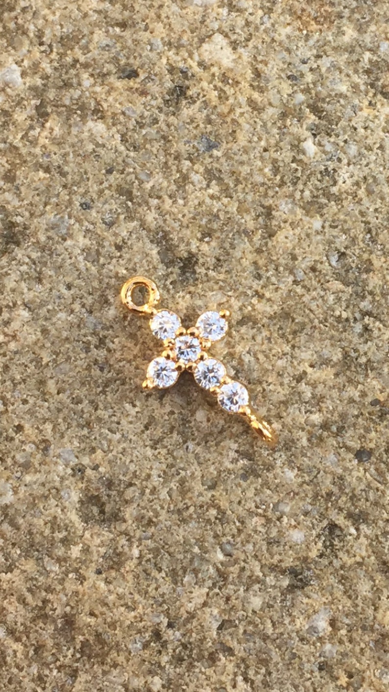 May include: Gold-tone cross charm with clear cubic zirconia stones. The charm has a loop at the top for attaching to a necklace or bracelet.