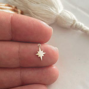 Peut inclure: Un petit charme en forme d'étoile de couleur or à huit branches, tenu entre les doigts. Le charme a une petite boucle en haut pour l'attacher à un collier ou un bracelet. Une gland blanc est en arrière-plan.