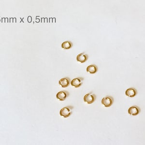 Può includere: Primo piano di 11 anelli di salto dorati, ognuno misura 2,5 mm x 0,5 mm. Gli anelli sono sparsi su una superficie bianca.