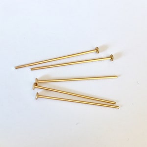 Peut inclure: Cinq épingles à tête ronde de couleur dorée. Les épingles sont disposées en éventail sur une surface blanche.